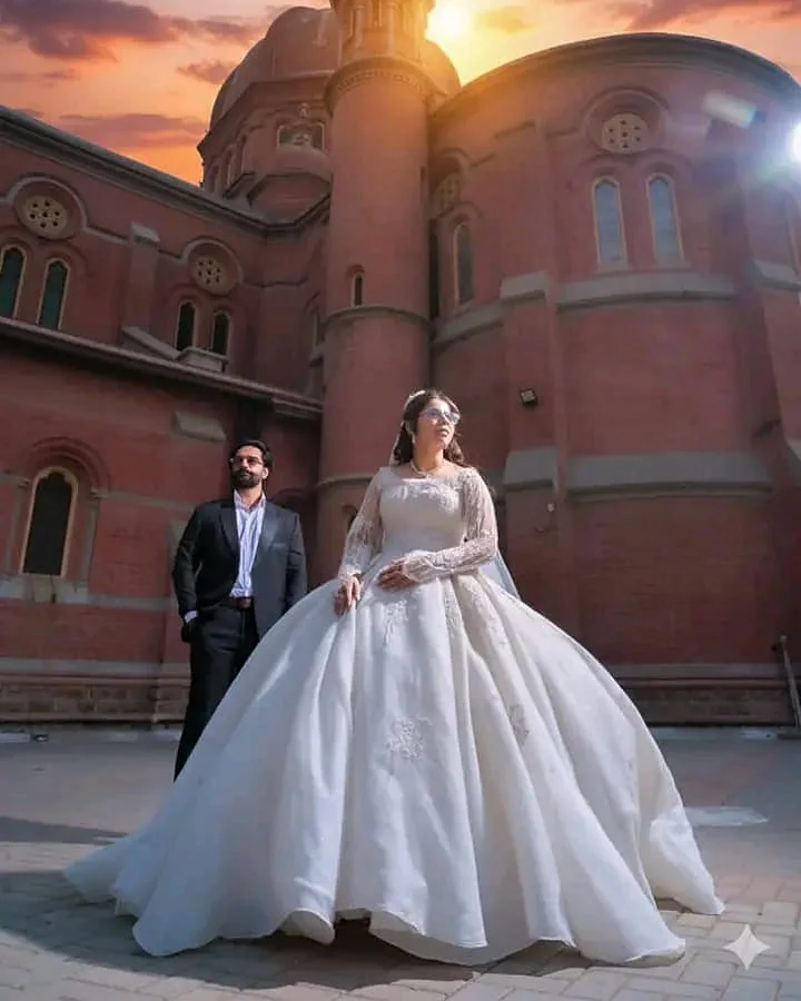 A bride in a white ball gown before a sunlit cathedral tower, with the groom standing behind her
