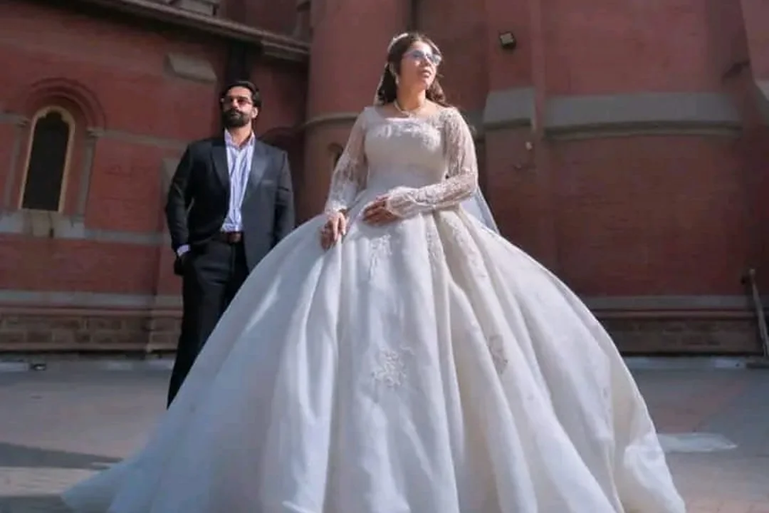 A bride in a full ball gown stands before a sunset-lit cathedral with the groom behind her